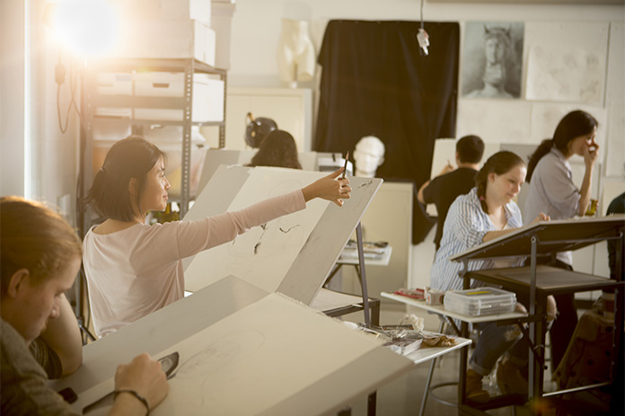 A student in Intermediate Drawing measures space with an outstretched arm and a pencil